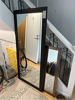 Three mirrors leaning against a wall and staircase. Largest has textured black frame; medium has metallic frame; smallest mirror's frame bent and damaged.