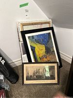 Stack of framed prints and canvas prints leaning against wall including a yellow building nighttime street scene and smaller framed print of ships in battle.