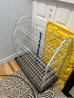 White wire 4-tier shoe rack with black plastic boot tray underneath and blue shoehorn attached, placed in corner on patterned rug near door.