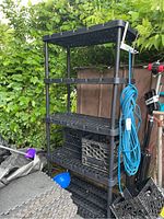 Black resin shelving unit with five shelves, shown outdoors with some cords and crates beside it, contents not included.