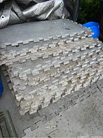 Stack of grey rubber interlocking floor tiles showing puzzle-like edges, some dirt and grime from outdoor storage.