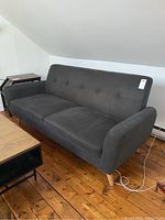 Front angled view of the dark grey three seater sofa showing the tufted back, armrests, and wood legs placed on wooden floor.