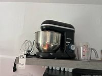 Full view of the Thinkkitchen stand mixer with stainless steel bowl and three attachments (dough hook, whisk, paddle) arranged on a shelf. Also shows two plastic liquid measuring cups next to the mixer.