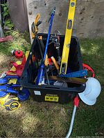 Black plastic tote filled with various hand tools including large yellow and small blue levels, white hard hat, red straps, and clamps.