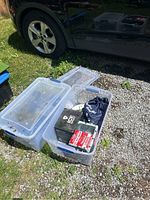 Two clear plastic totes outdoors containing various automotive tools and accessories. One tote is open and shows a hydraulic trolley jack, axle stands, and a torque wrench case inside. The other tote contains black bags, epoxy boxes and a hole saw.