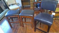 Three bar stools side by side. Two stools have square, backless espresso-colored seats with silver nailhead trim and wooden legs with horizontal footrests. One stool has an espresso-colored padded backrest and seat, no nailhead trim visible.