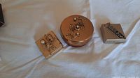 Three vintage metal pill boxes arranged on white cloth: round copper-tone floral, square gold-tone floral with a pearl, and small gold-tone textured with zipper pill holder