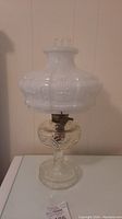 Full view of antique kerosene lamp showing white embossed milk glass shade and clear etched glass base on white surface.