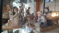 Photo showing four assorted porcelain figurines displayed on glass shelf with reflections, including children on a sled and couple figurines.