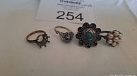 Four assorted ladies silver rings displayed on a white surface: one with empty setting, one with dark stone, one with turquoise and floral design, one with pearls cluster.