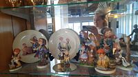 Shelf display showing 8 Hummel figurines and 2 Hummel plates in the background.