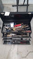 Top lid open showing assorted hand tools including pliers, wrenches, screwdrivers, and more inside the toolbox.