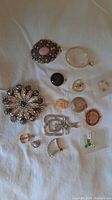 Photo showing an assortment of vintage and possibly antique necklace pendants and charms arranged on fabric background. Includes a large silver floral medallion, pink stone oval pendant, magnifying glass pendant, and other small charms.