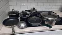 Photo showing assorted pots and pans with lids arranged on a kitchen counter including frying pan, sauté pan, saucepans, stockpot, and metal lids.