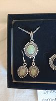 Close-up photo showing silver necklace with oval green stone pendant and matching drop earrings with oval green stones in silver frames.