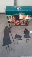 Black metal yard art Nativity figures including Joseph, Mary with baby Jesus in manger, all with ground stakes for outdoor display. Original boxed vintage Grandeur Noel 9-piece porcelain Nativity shown in background.