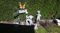 Several pieces of yard art displayed on grass including a topiary with butterfly and welcome sign, concrete statues (child and dog), and smaller decorative animals.