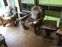 Two solid wood armchairs with wide flat armrests and country cottage scenic upholstery next to matching wooden side table. Floor plywood visible.