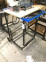 Front side view of Mastercraft power tool stand with wooden mounting surface and wheels on black metal frame with blue adjustment bars.