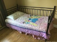 Side view of black metal bed frame, mattress and Disney Princess bedding with pillow on wooden floor next to window.