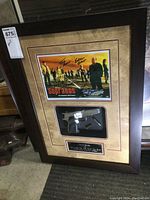 Framed piece with signed poster mounted above plastic replica gun and plaque