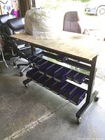 View of metal work bench showing MDF top, metal frame, wheels, and blue plastic storage bins underneath from a side angle.