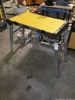 Work stand fully set up, showing yellow plastic work surface and metal legs with wheels on one side.