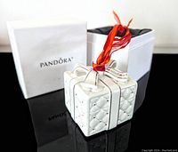 Front and side angle of the white ceramic Pandora 2016 Christmas ornament designed as a quilted present with silver highlights and a red ribbon, alongside its original white box.