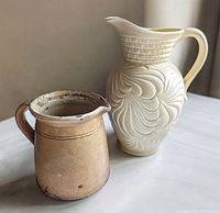 Photo showing both antique jugs together on a light surface. The smaller brown glazed earthenware jug is on the left, and the taller cream-colored Art Nouveau ceramic pitcher is on the right.