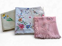 Three folded vintage embroidered linens: natural with floral embroidery, white heavily embroidered square, and pink fringed linen with musical embroidery.