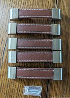 Top view of 5 brown leather and brushed silver metal cabinet pulls arranged on wood surface with screws below