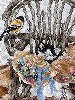Close-up photo showing detailed cross stitch of a wooden chair with a yellow bird on the back and a sleeping black and white cat on the seat, alongside a floral hat with a blue ribbon.