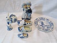Photo of seven Delft blue porcelain pieces including wall plate, three shoe ashtrays, figurine, bunny bank, and bell on white background