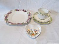 Photo of all four porcelain items displayed together: teacup with saucer and biscuit plate behind it, square handled serving plate to the left, heart shaped mint dish in front.