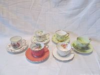 Six assorted John Aynsley & Sons teacup and saucer sets displayed, each with unique patterns and certain matching saucer colors such as green, coral, white, and floral designs.