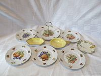 Full set of nine porcelain fruit-pattern dishes and bowls arranged together on a white background, showing the variety of fruit designs and styles including handled serving dishes and lusterware bowls.