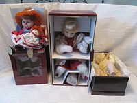 Three porcelain dolls arranged: red-haired Elby Musical doll seated on original box, blonde Ashton Drake doll in box with padding, and another blonde Artisan Collection doll in box.
