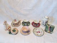 Full view of entire lot including the nine porcelain items arranged with a light backdrop showing wall pocket, coffee pot, teacups and saucers, saucer, figurine, and planter.