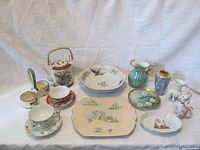 Photo showing the full lot of 12 Japanese porcelain items arranged on a white cloth, includes teacups, pitchers, vases, serving dishes, and figurine.