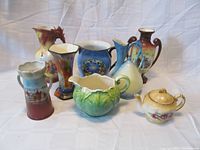 Group photo showing all eight porcelain items including vases and pitchers with varied pastel colors and hand-painted scenes, plus the teapot in front.