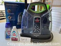 Bissell SpotClean handheld cleaner with attached hose, two cleaning formula bottles, and original box in background.