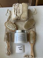Top view showing the SkinIn body butter jar, four wooden brushes with different bristle types, three beige exfoliating bath scrubs, and a beige drawstring pouch all on a white surface.