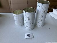 Three cylindrical glass vases of varying sizes decorated with gold and silver stars and snowflakes. Each vase contains a fairy light system inside to light up the vase. Vases are brand new, batteries not included.
