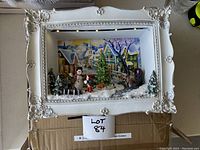Front view of illuminated white ornate shadow box showcasing a snowy Christmas village scene with multiple miniature figurines including a snowman, children, Christmas tree, and pine trees.