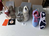 Three pairs of footwear displayed side by side: brown and white leather sandals, white running sneakers with Velcro straps, and blue/pink running sneakers with laces, with Vionic shoeboxes in background.