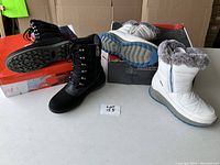 Pair of new black and white leather winter boots placed on and next to their boxes. Black boots have laces and white boots have faux fur trim.