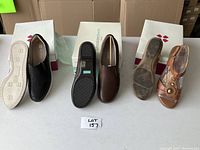 Lot of three shoes displayed on white table with shoeboxes behind: one black slip-on loafer, one brown slip-on loafer, one tan sandal.