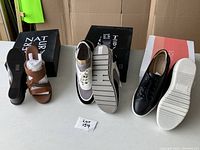 Photo showing all three pairs of shoes with their accompanying Nat-2 branded boxes: brown leather sandals, black leather sneakers, and grey/white/black runners.