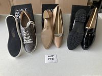 Three pairs of women's shoes arranged side by side: silver tinted running sneakers on left, beige low heels center, black patent leather slip-ons right, with boxes behind them.