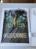 Full view of the solar powered butterfly welcome sign wrapped in protective plastic, showing the colorful painted butterfly and the white 'WELCOME' text on the black base panel.